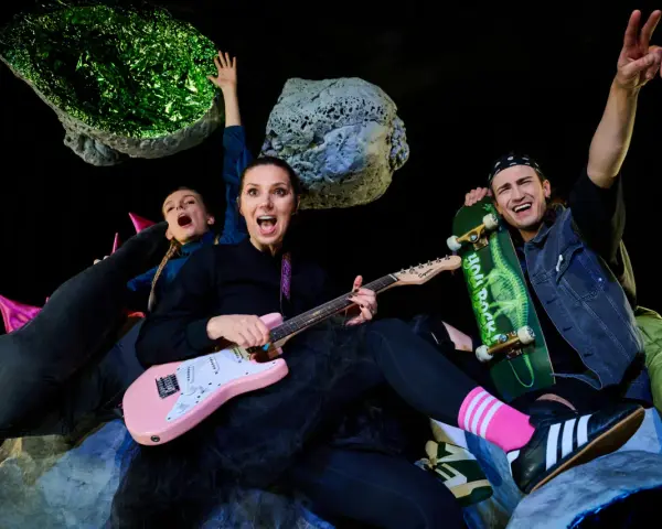 Energiczna ekipa w kosmicznej scenografii, grająca polski folk rock. Kobieta z różową gitarą, mężczyzna ze skateboardem, wesołe miny i podniesione ręce.