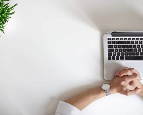 Minimalistyczne biurko z laptopem, zieloną rośliną i dłońmi złożonymi w geście oczekiwania. Idealne do wizualizacji pracy kreatywnej i renderowania.