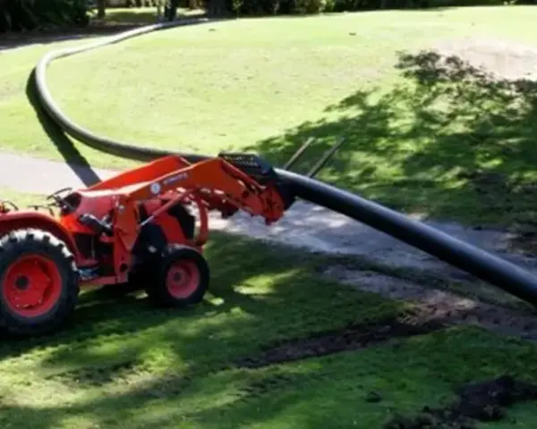A tractor with a front loader is laying a long, flexible HDPE pipe across a grassy area.