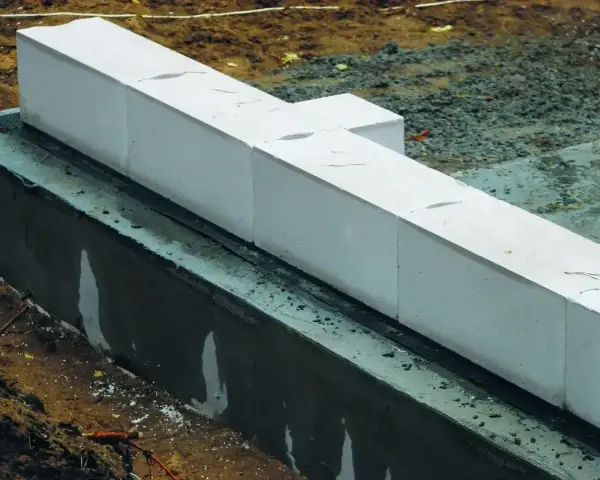 Białe bloczki betonowe układane na warstwie izolacji. Widać wilgotną zaprawę i ziemię wokół fundamentów.