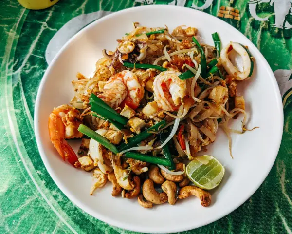 Talerz Pad Thai z krewetkami, kalmarami, kiełkami i orzechami nerkowca, udekorowany ćwiartką limonki.