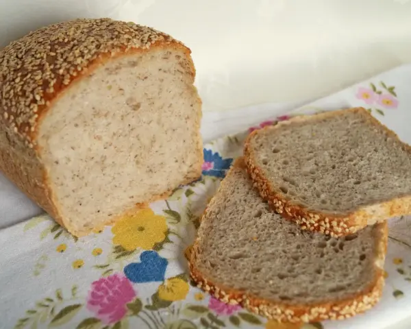 Chleb z kaszą gryczaną, posypany sezamem, leży obok dwóch kromek na kwiecistym obrusie.