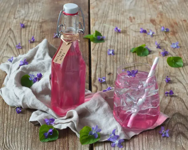 Flasche mit Veilchensirup und ein Glas mit Eis und Veilchenblüten auf Holzhintergrund.