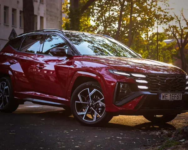 Czerwony Hyundai Tucson Hybrid zaparkowany na tle jesiennych drzew i kamienicy.