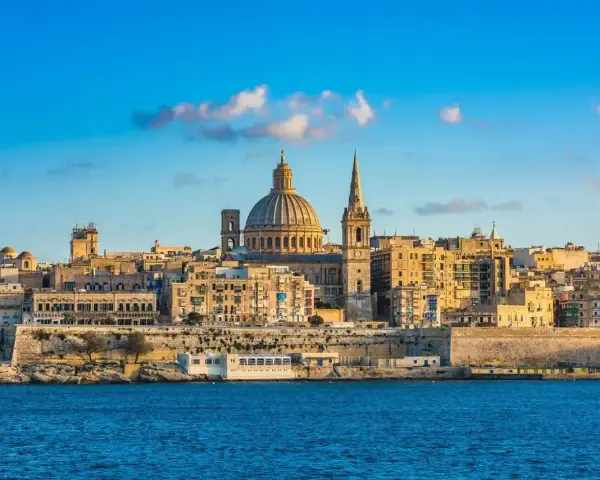 Malta, wyspa do kogo należy? Widok na Vallettę z kopułą i wieżą kościelną, otoczoną zabytkową architekturą, nad błękitnym morzem.