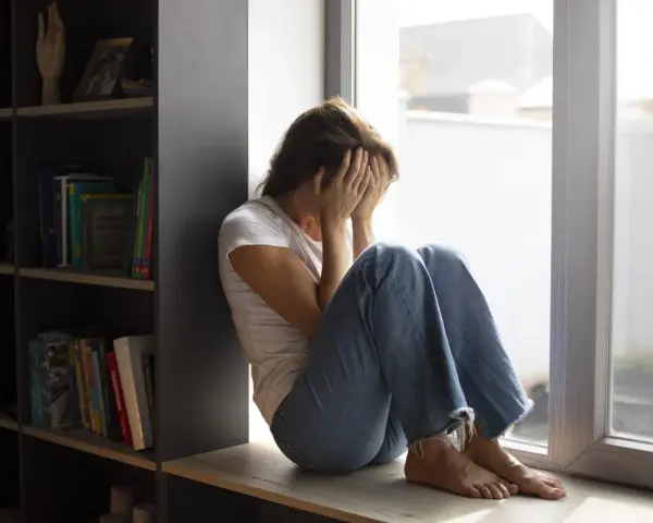 Kobieta siedzi przy oknie, zakrywając twarz dłońmi. Wokół panuje nastrój melancholii, odzwierciedlający dystymię.