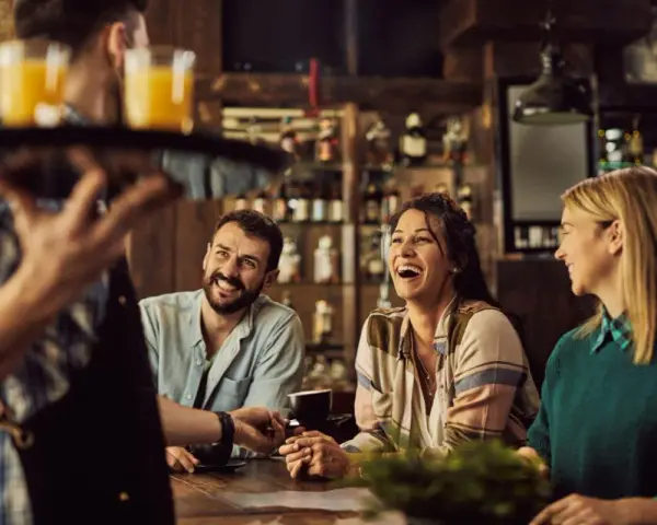 Un camarero sirve bebidas a un grupo de amigos riendo en un bar. La conversación animada sugiere que aún no es tarde para el ruido.