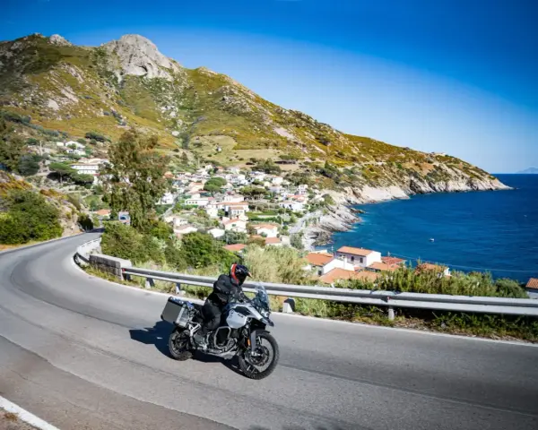 Un motociclista esplora le curve panoramiche dell'Elba, con il mare blu e un villaggio pittoresco sullo sfondo.