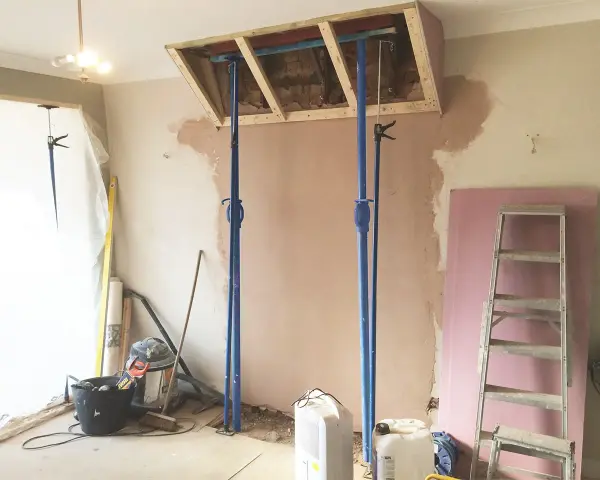 Travaux de rénovation : des étais soutiennent une structure en bois au-dessus d'une zone où l'on s'apprête à casser une cheminée. Matériaux de construction et outils au sol.