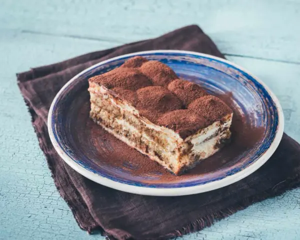 Pyszny kawałek prawdziwego tiramisu, posypany kakao, na niebieskim talerzu.