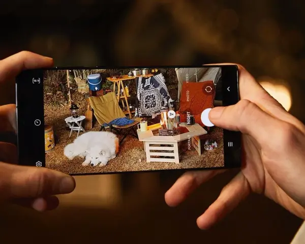 Une photo de téléphone capture un chien blanc endormi lors d'un camping. L'écran du téléphone montre une scène de camping avec une tente, une chaise pliante et un feu de camp.
