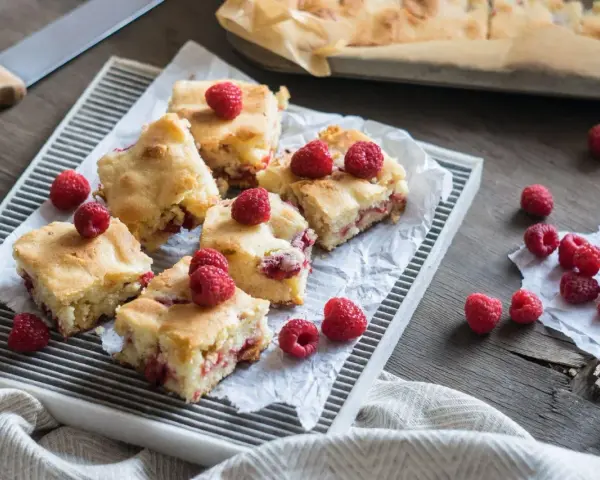 Słodkie brownie i blondie z malinami, pokrojone na kwadraty, ułożone na tacy. Kilka malin rozsypanych obok.