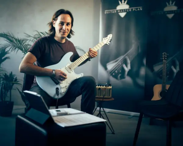 Mann mit E-Gitarre, bereit, online Gitarre zu lernen. Noten und Tablet liegen bereit.