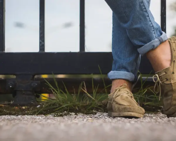 Gambe con jeans arrotolati e scarpe senza calzini, appoggiate su un muretto con erba.