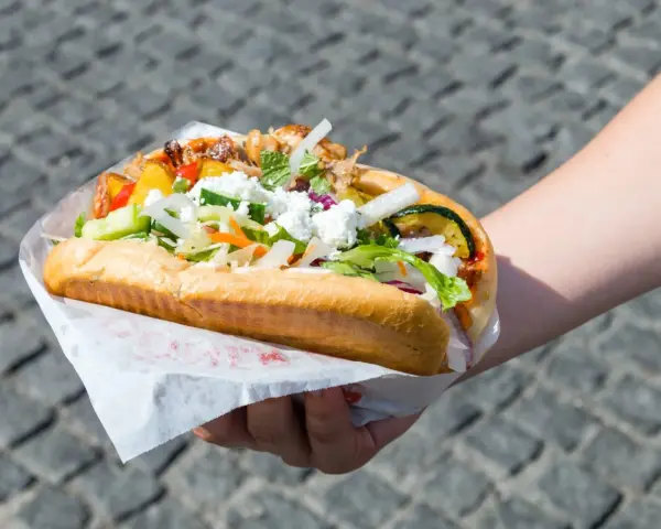 Kebab w bułce z mięsem, warzywami i serem feta, trzymany w dłoni.
