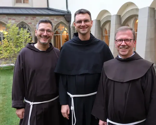 Drei Franziskaner Minoriten in braunen und schwarzen Kutten lächeln in die Kamera vor einer Klosterfassade.