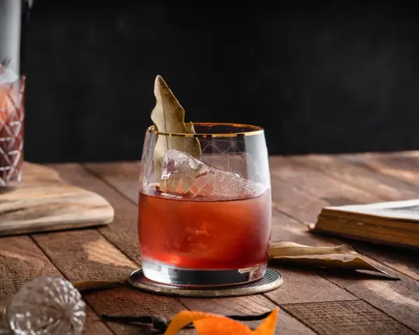 Czerwony koktajl z lodem i liściem laurowym w szkle, obok karafki i starej książki. To może być drink z wermutem.