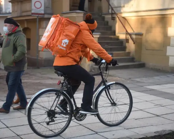 Kurier rowerowy w pomarańczowej kurtce z plecakiem Pyszne.pl, zarobki zależą od liczby dostaw.