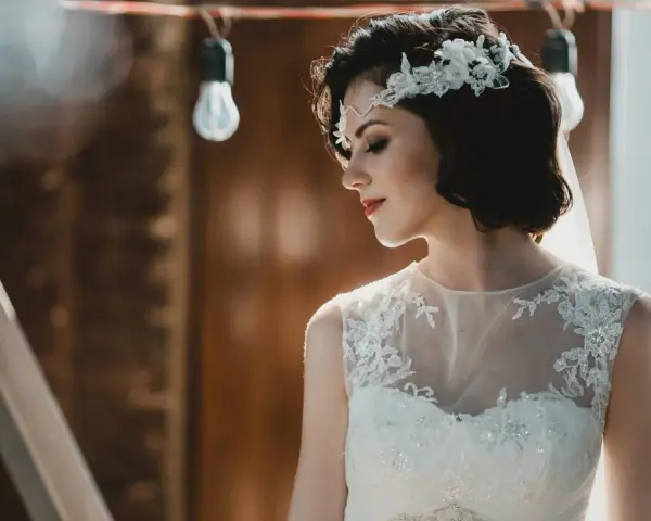 Panna młoda z krótkimi włosami w stylu boho, ozdobionymi kwiatową opaską, w białej sukni z koronkowymi detalami.