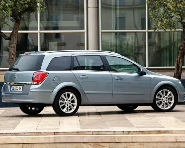 Srebrny Opel Astra H kombi zaparkowany przed nowoczesnym budynkiem z drzewami.
