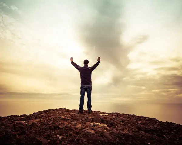Mann mit erhobenen Armen auf einem Felsen, blickt auf das Meer. Ein Gefühl des aaronitischen Segens liegt in der Luft.