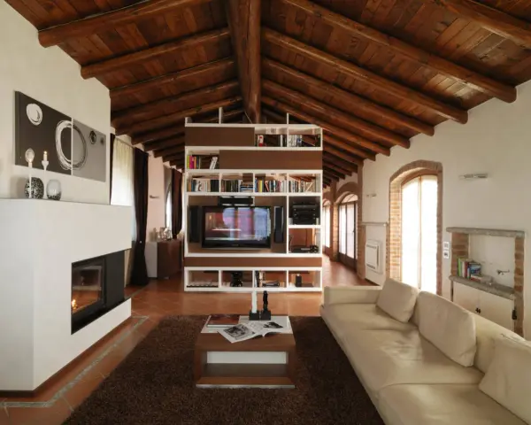 Accogliente soggiorno mansardato con soffitto in legno a vista, camino moderno e libreria centrale. Idee mansarde in legno per un ambiente caldo.