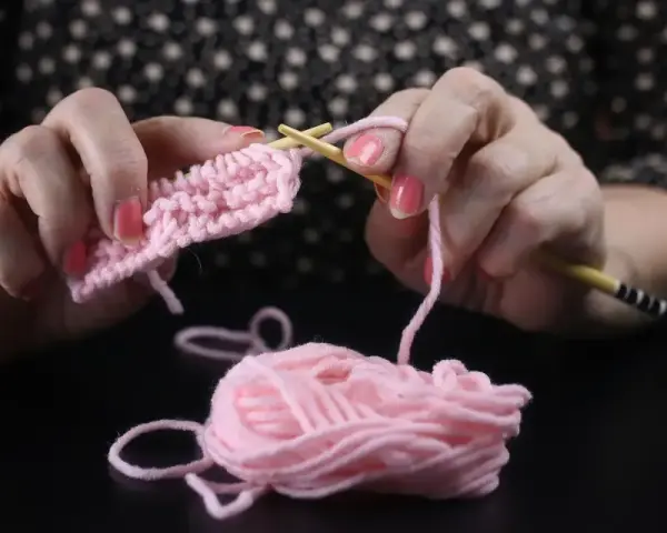 Händerna stickar en rosa v-ringning med ljusa trästickor. En garnnystan ligger bredvid.