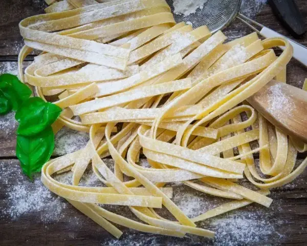 Świeży, domowy makaron bez glutenu, posypany mąką, z listkami bazylii i drewnianą łyżką.