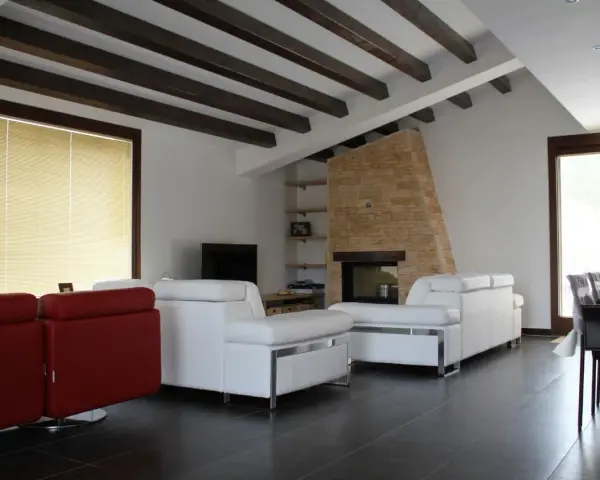 Soffitto con travi in legno scuro, un camino in pietra e divani bianchi. Ecco come fissare travi in legno al soffitto per un effetto rustico.