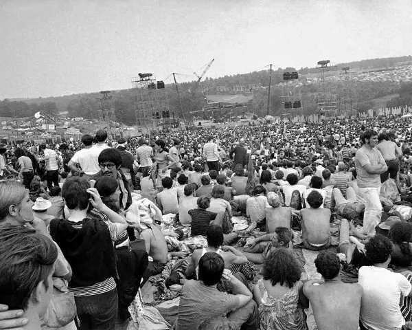 Tysiące ludzi siedzi na trawie, patrząc na scenę festiwalu. To jest Woodstock, gdzie jest muzyka i wolność.