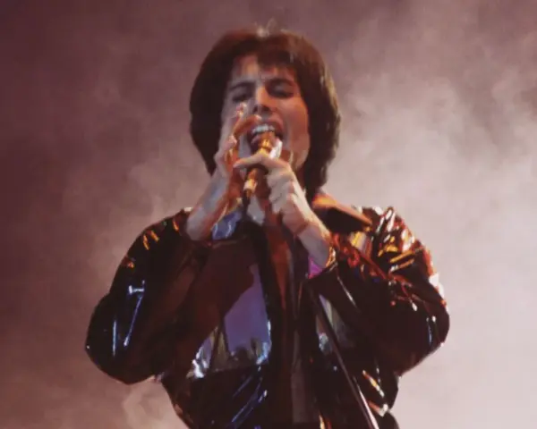 Freddie Mercury śpiewa z pasją, czując, że chce się uwolnić. Błyszcząca kurtka odbija światła sceny.