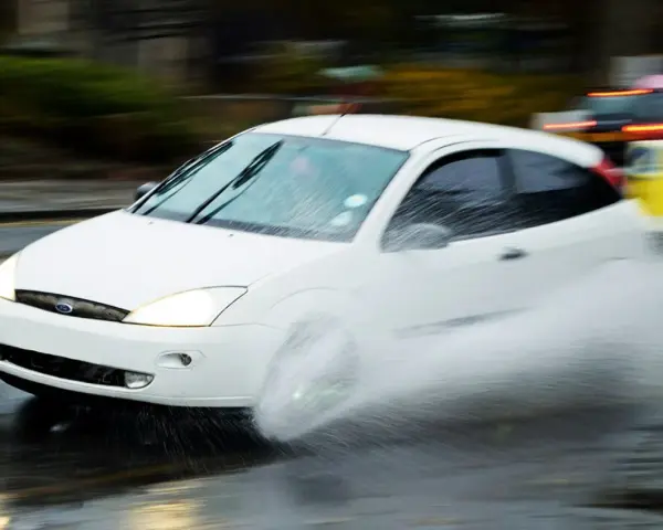 Auto bianco in movimento su strada bagnata, con un'enorme spruzzata d'acqua che indica l'aquaplaning cos'è.