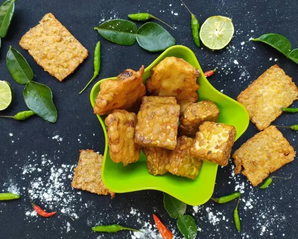 Knusprig gebratener Tempeh, ein fermentiertes Sojaprodukt, liegt in einer grünen Schale. Dazu garnieren Limettenscheiben, grüne Chilis und Blätter.