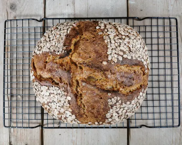 Ein rustikales Porridge Brot mit Haferflockenkruste, das auf einem schwarzen Gitter auf Holzdielen abkühlt.