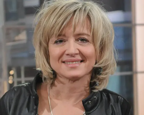 Uśmiechnięta Małgorzata Bogdańska w czarnej ramonesce, z blond włosami i delikatnym makijażem.