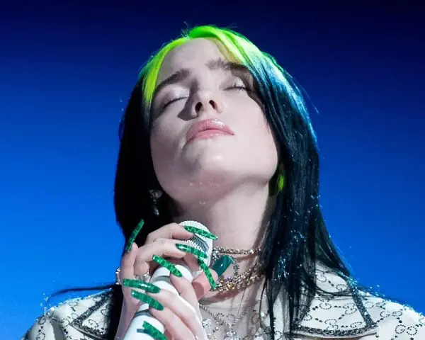 Piosenkarka Billie Eilish z zamkniętymi oczami śpiewa do mikrofonu, jej zielone pasemka włosów lśnią.