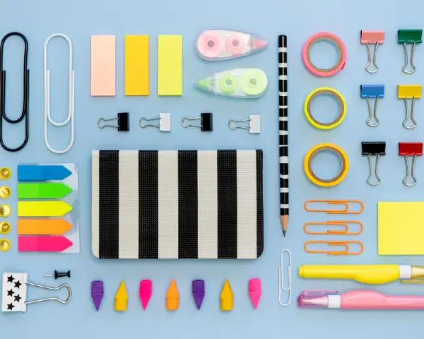 Un surtido vibrante de material de oficina lista para usar: clips, notas adhesivas, cintas, lápices y más, organizados sobre un fondo azul claro.