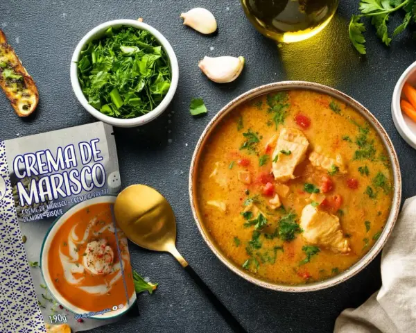 Un tazón de sopa de pescado casera con trozos de pescado, tomates y perejil, junto a una tostada, zanahorias y un paquete de crema de marisco.