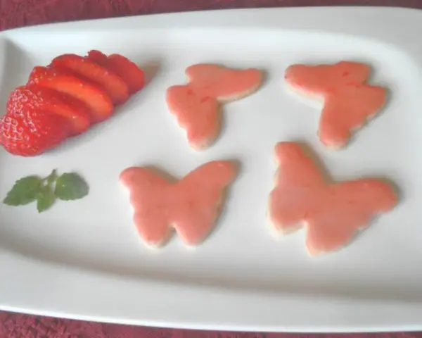 Zarte Erdbeer-Kekse in Schmetterlingsform, dekoriert mit rosa Zuckerguss, neben frischen Erdbeerscheiben und einem Minzblatt.