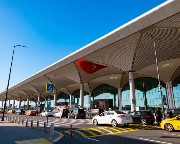 Nowoczesny budynek lotniska Bodrum z charakterystycznym zadaszeniem. Przed terminalem ruch samochodowy, w tym żółte taksówki.