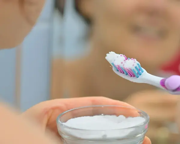 Ręka trzymająca szczoteczkę do zębów z białym proszkiem, obok miseczka z sodą oczyszczoną. Domowa pasta do zębów gotowa.