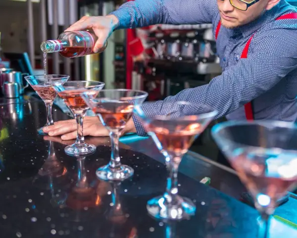 Barman nalewa różowy drink do kieliszków koktajlowych na czarnym blacie.