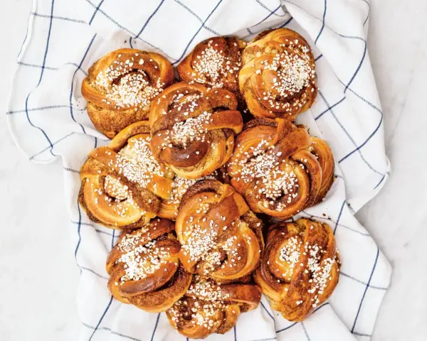 En hög med gyllene kanelbullar, toppade med pärlsocker, ligger på en vit handduk med blå rutor. Camilla Hamid skulle vara stolt över dessa kanelbullar.