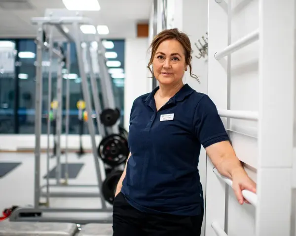 En kvinna i träningskläder står i ett gym. Hon har en namnskylt på sin tröja. I bakgrunden syns träningsredskap, som kan användas för att stärka underbenets muskler.