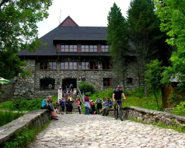 Kamienny budynek schroniska w Dolinie Chochołowskiej z ludźmi na schodach i rowerzystą na kamiennej drodze.