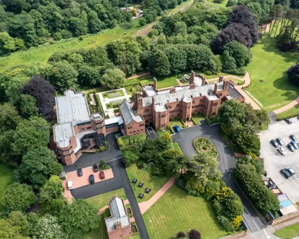 Aerial view of the abbey guest house, a grand brick building surrounded by lush green trees and manicured lawns. Cars are parked in a lot.