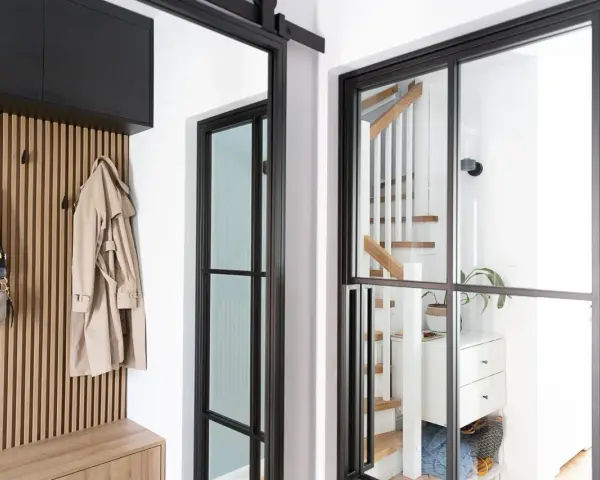 Nowoczesny wiatrołap z drewnianą boazerią, czarnymi przeszkleniami i widocznymi schodami. Na wieszaku płaszcz i torebka.