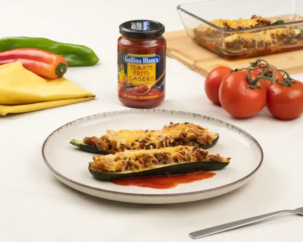 Dos calabacines rellenos de carne y gratinados con queso, servidos en un plato con tomate frito.