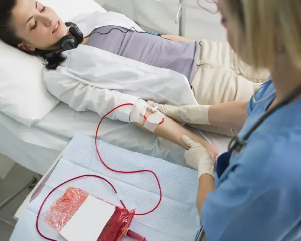 Kobieta leży w łóżku szpitalnym, słuchając muzyki przez słuchawki. Pielęgniarka wykonuje przetaczanie krwi, podłączając worek z krwią do jej ręki.