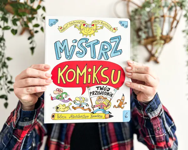 Okładka książki "Mistrz Komiksu" z rysunkami postaci i słońca, trzymana przez ręce.